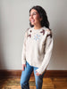 A woman wearing a cream-colored knit sweater with brown and light blue floral details on the shoulders and chest, paired with blue jeans, standing in a room with a neutral wall and wooden floor.
