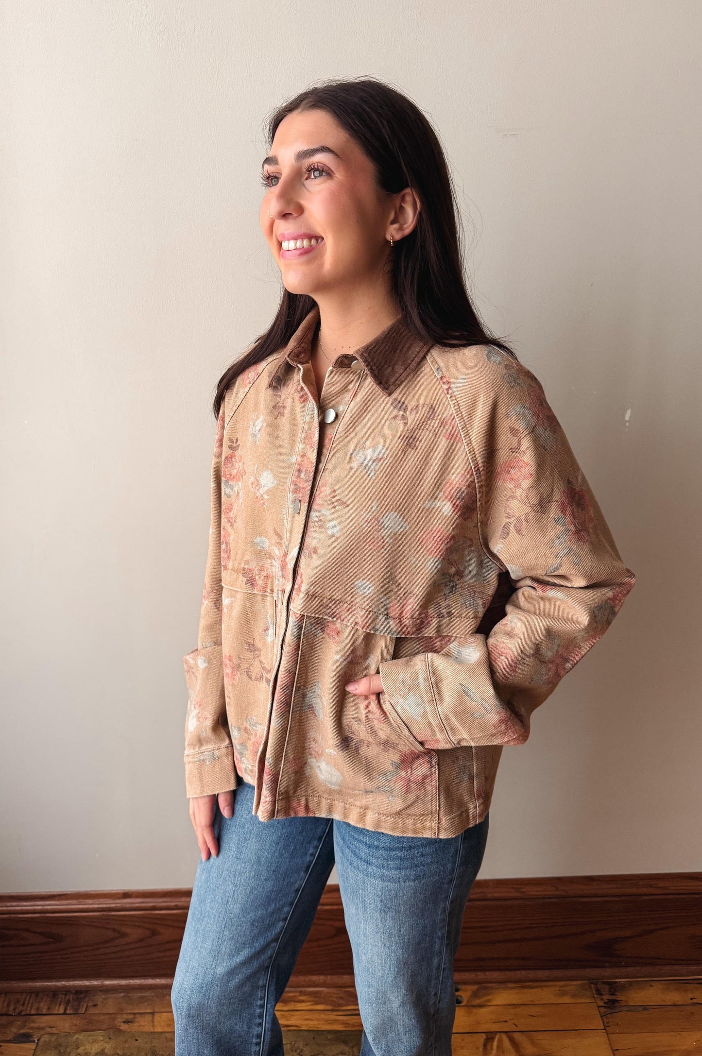 A woman wearing a beige button-up shirt jacket with a pink and white floral pattern, paired with blue jeans, standing against a plain beige wall.