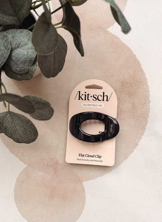 A black flat cloud clip by kit-sch on its original beige cardboard hanger, styled on a beige patterned surface with green eucalyptus leaves.