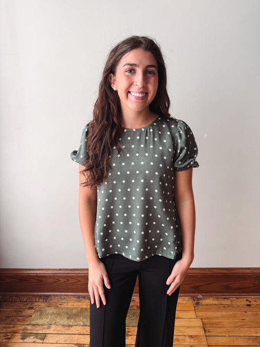 A smiling young woman with long brown hair stands indoors wearing a sage green short-sleeve blouse with white polka dots and black pants, set against a plain off-white wall and wooden floor.