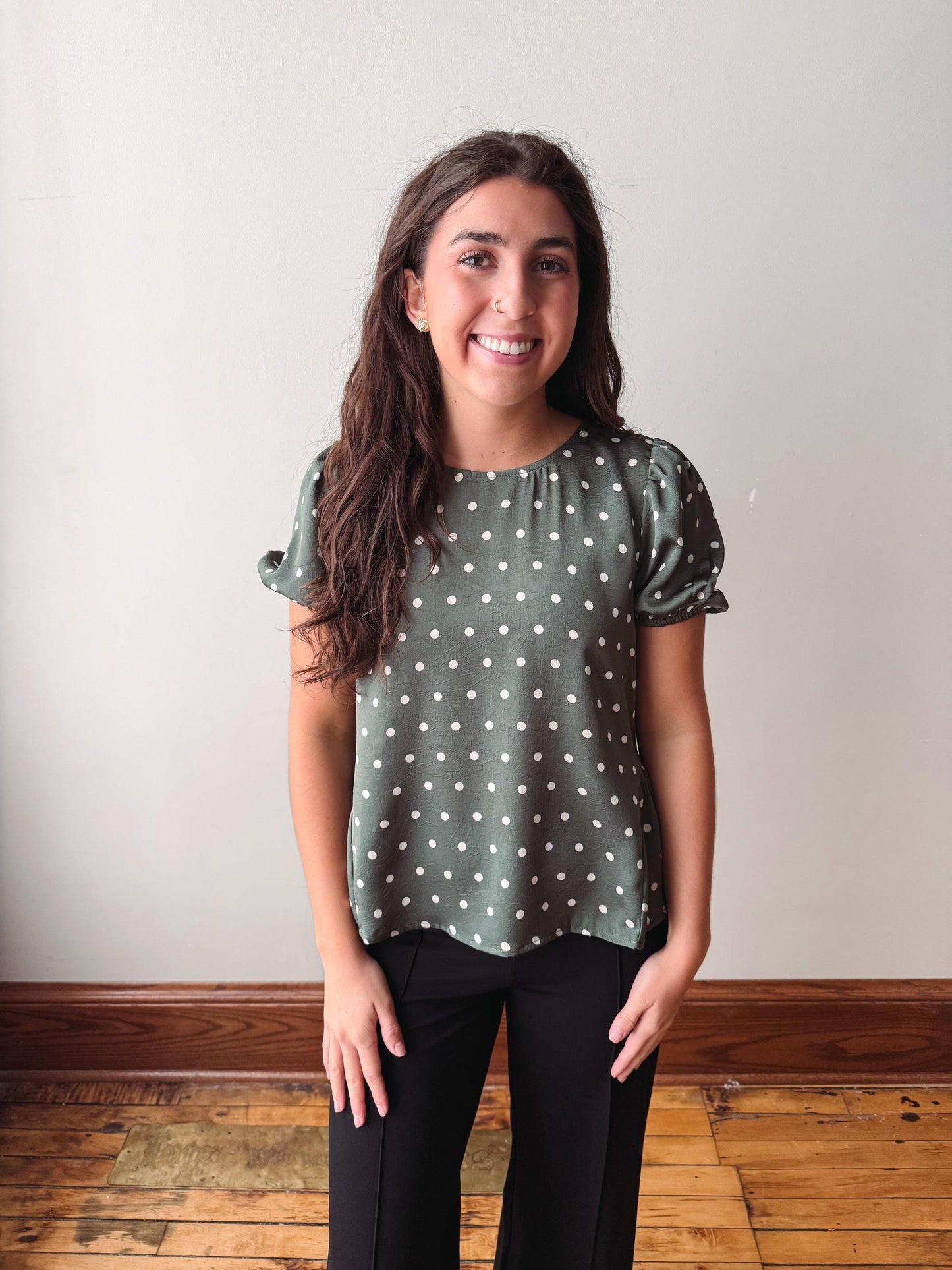A smiling young woman with long brown hair stands indoors wearing a sage green short-sleeve blouse with white polka dots and black pants, set against a plain off-white wall and wooden floor.