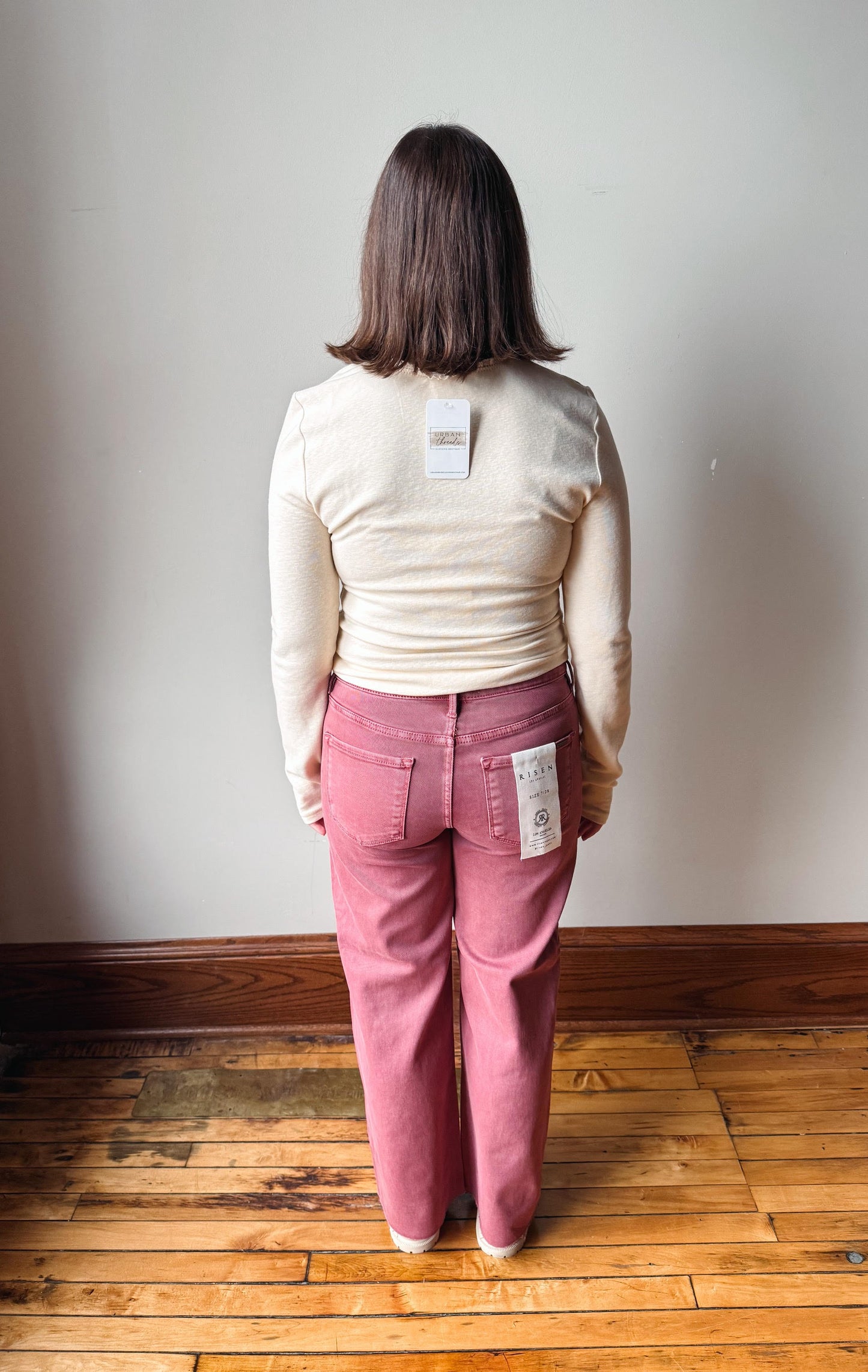 A woman viewed from behind wearing a cream ribbed long-sleeve top and dusty rose pink wide-leg jeans, standing on a wooden floor against a plain off-white wall.