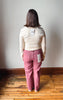 A woman viewed from behind wearing a cream ribbed long-sleeve top and dusty rose pink wide-leg jeans, standing on a wooden floor against a plain off-white wall.