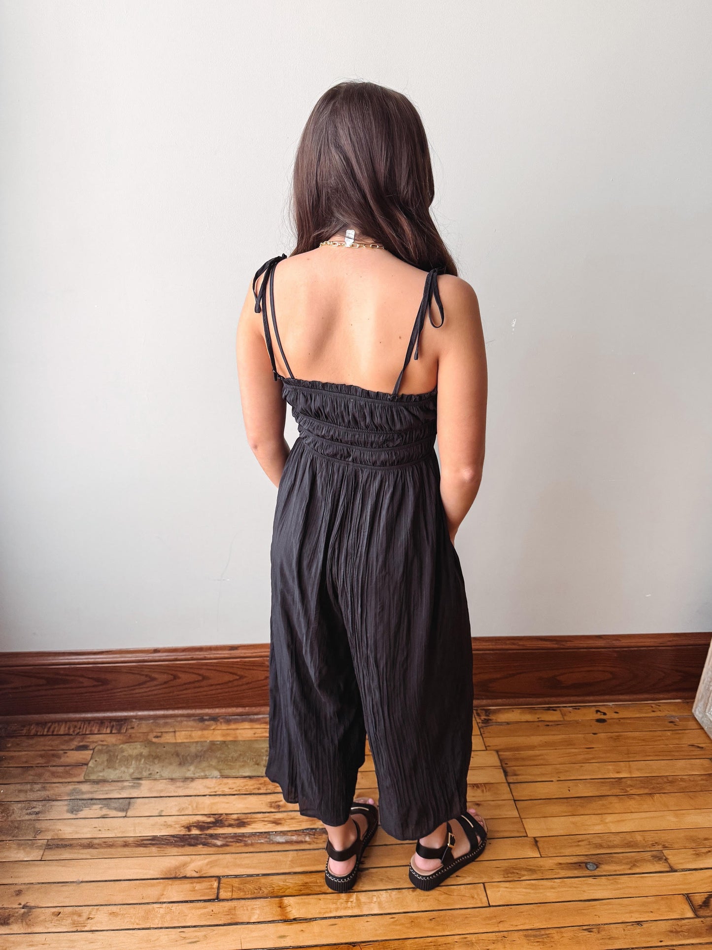 Person wearing a black jumpsuit standing on a wooden floor with a white wall background