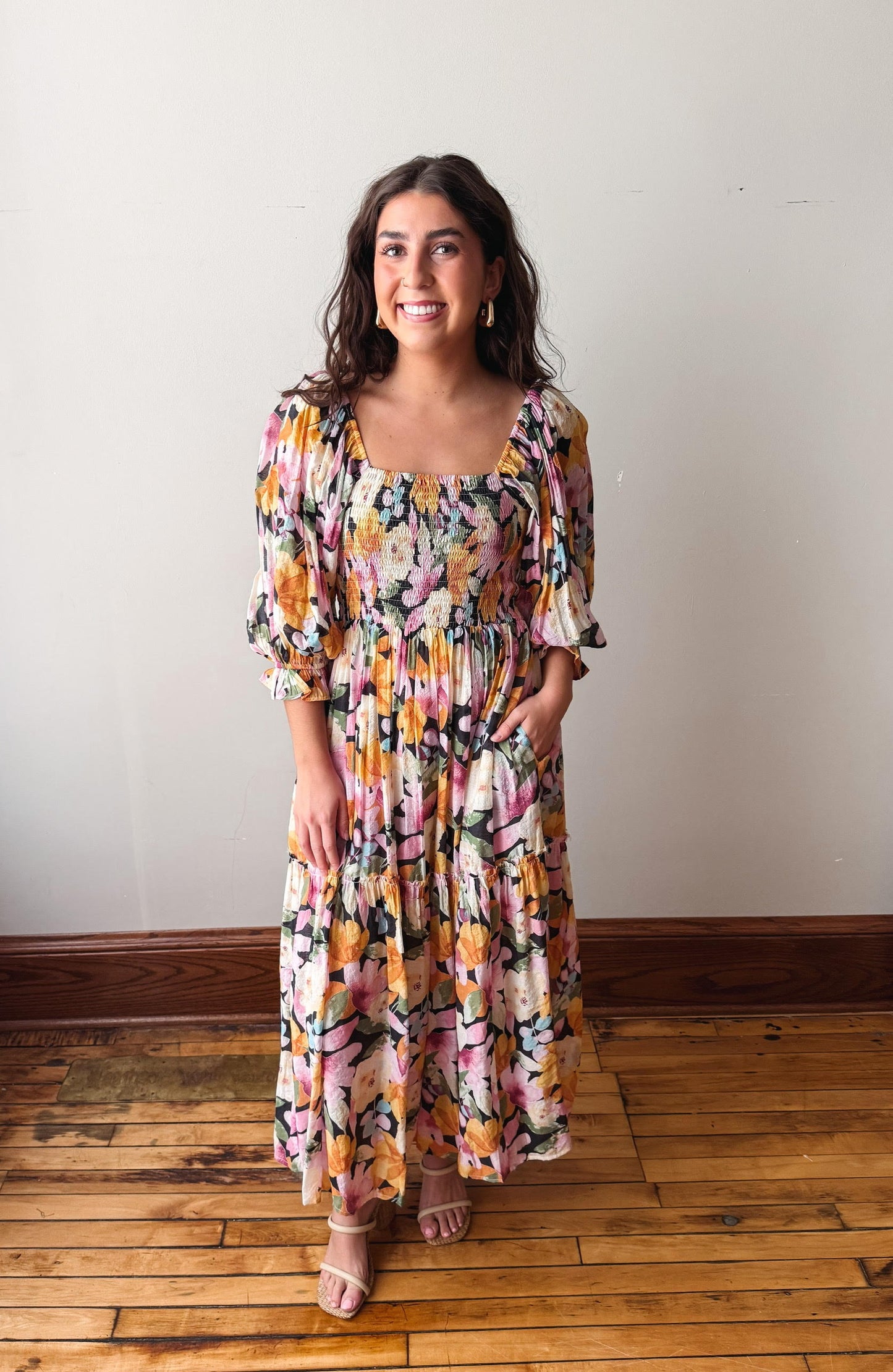 Woman wearing a colorful floral dress standing on a wooden floor with a white wall background