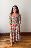 Woman wearing a colorful floral dress standing on a wooden floor with a white wall background