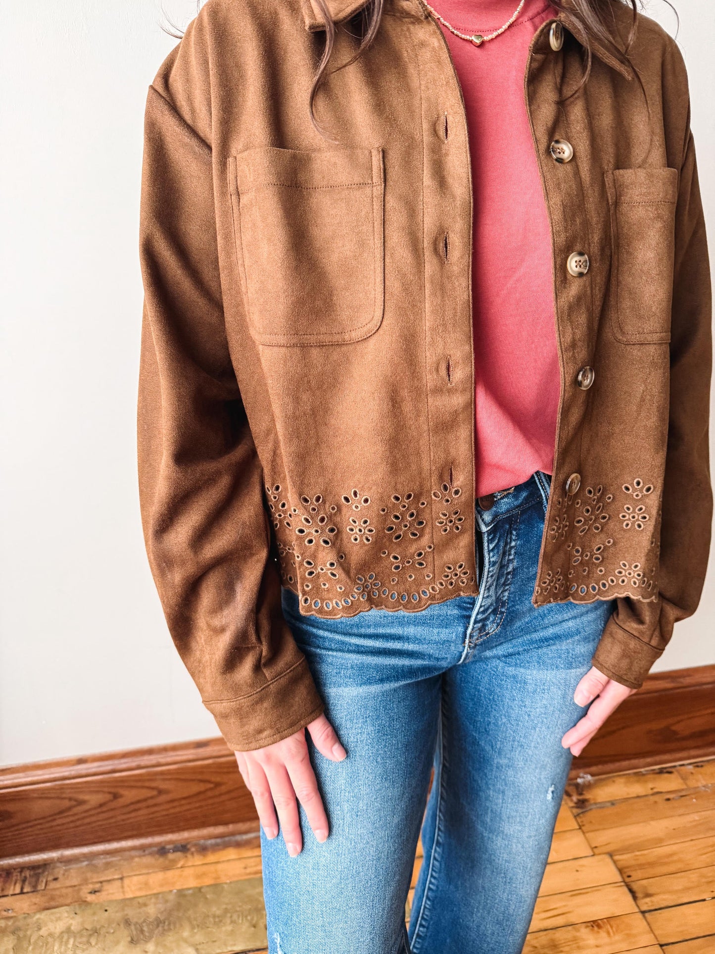 A person wearing a brown suede-like button-front jacket with decorative eyelet embroidery near the hem, layered over a pink top and blue jeans, standing indoors on a wood floor.