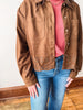 A person wearing a brown suede-like button-front jacket with decorative eyelet embroidery near the hem, layered over a pink top and blue jeans, standing indoors on a wood floor.