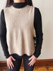 A person wearing a beige ribbed knit sweater vest over a black long-sleeve mock neck top and black pants, standing indoors against a light wall.