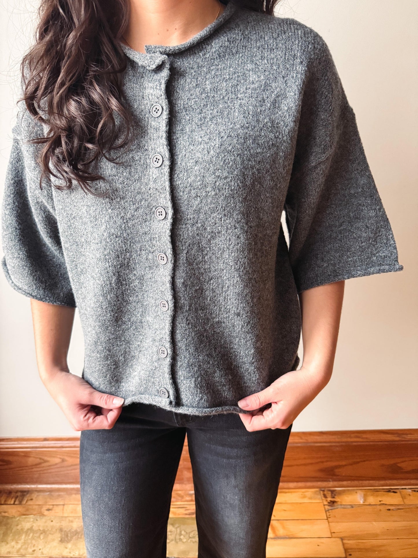 A woman wearing a gray knit short-sleeve button-front cardigan with dark gray jeans, standing indoors against a beige wall and wooden floor.