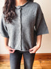 A woman wearing a gray knit short-sleeve button-front cardigan with dark gray jeans, standing indoors against a beige wall and wooden floor.