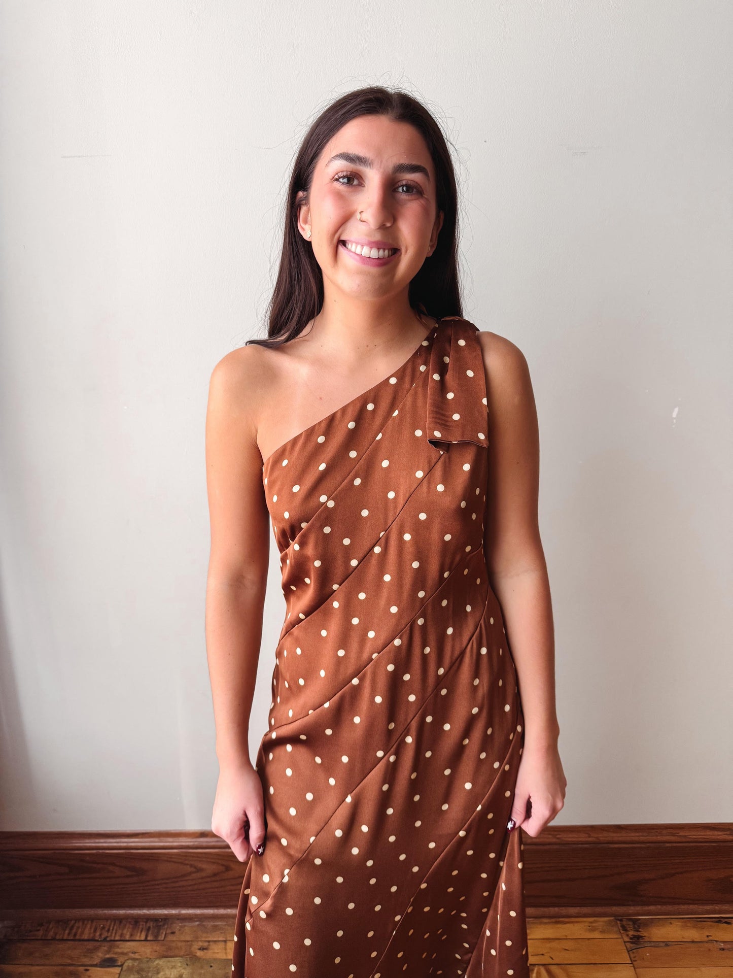A woman wearing a brown one-shoulder dress with white polka dots, standing against a plain off-white wall with a wooden baseboard and floor visible.
