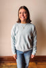 A smiling woman with shoulder-length brown hair stands indoors wearing a light heather gray crewneck sweatshirt and blue jeans against a plain beige wall with a wooden baseboard.