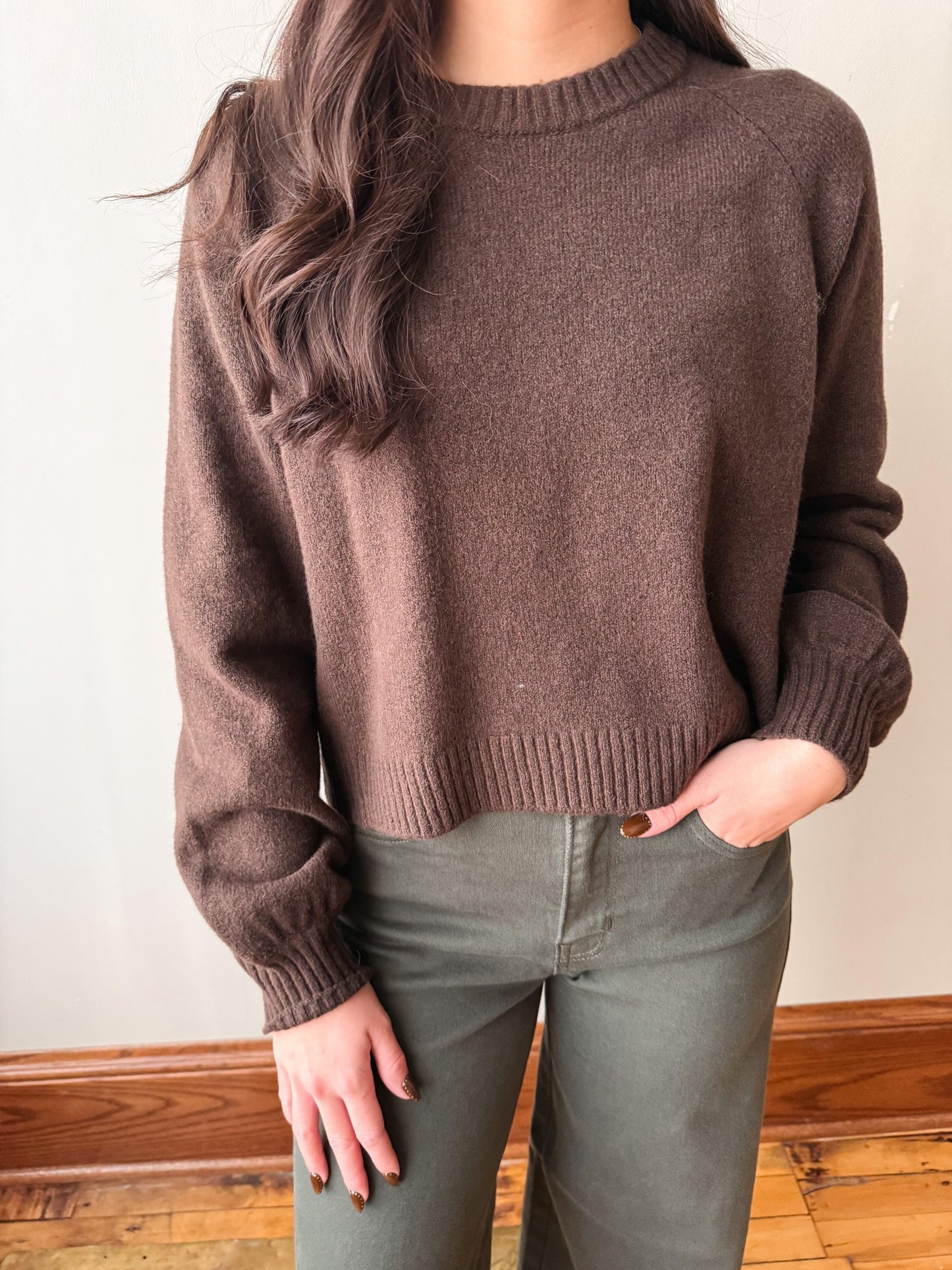 A woman wearing a brown knit crewneck sweater and olive green pants, standing indoors against a light wall with wooden trim and floorboards.