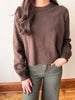 A woman wearing a brown knit crewneck sweater and olive green pants, standing indoors against a light wall with wooden trim and floorboards.