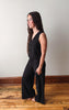Woman wearing a black sleeveless top and wide-leg pants standing on a wooden floor against a white wall.