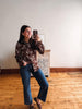 A woman takes a mirror selfie in a room with hardwood floors and a white wall, wearing a brown long-sleeve button-up shirt with a white abstract pattern, blue jeans, and brown flats.