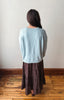 A woman is shown from behind wearing a light blue knit sweater and a dark brown patterned skirt, standing on a wooden floor against a plain off-white wall.