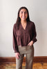 A woman modeling a dark brown long-sleeve blouse with a lace V-neck bodice and button front, paired with khaki pants, standing against a plain off-white wall.