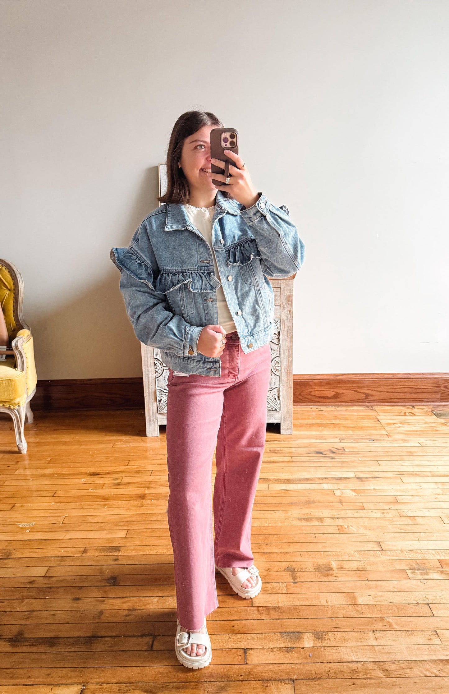 A woman takes a mirror selfie in a room with wooden floors and a white wall, wearing a light blue ruffled denim jacket, a white top, pink wide-leg pants, and white slide sandals.