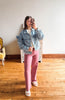 A woman takes a mirror selfie in a room with wooden floors and a white wall, wearing a light blue ruffled denim jacket, a white top, pink wide-leg pants, and white slide sandals.
