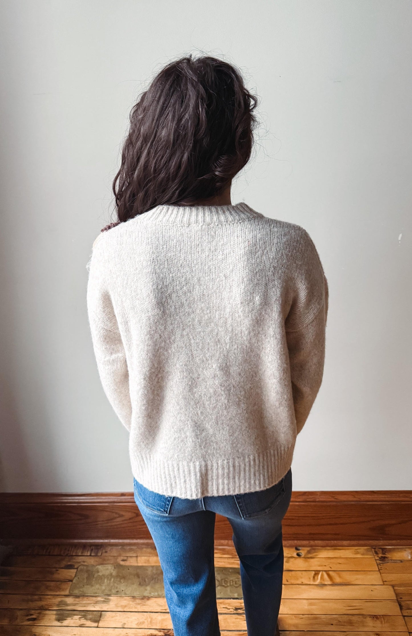 A person viewed from behind wearing a cream-colored knit sweater and blue jeans, standing on a wooden floor against a plain light wall.