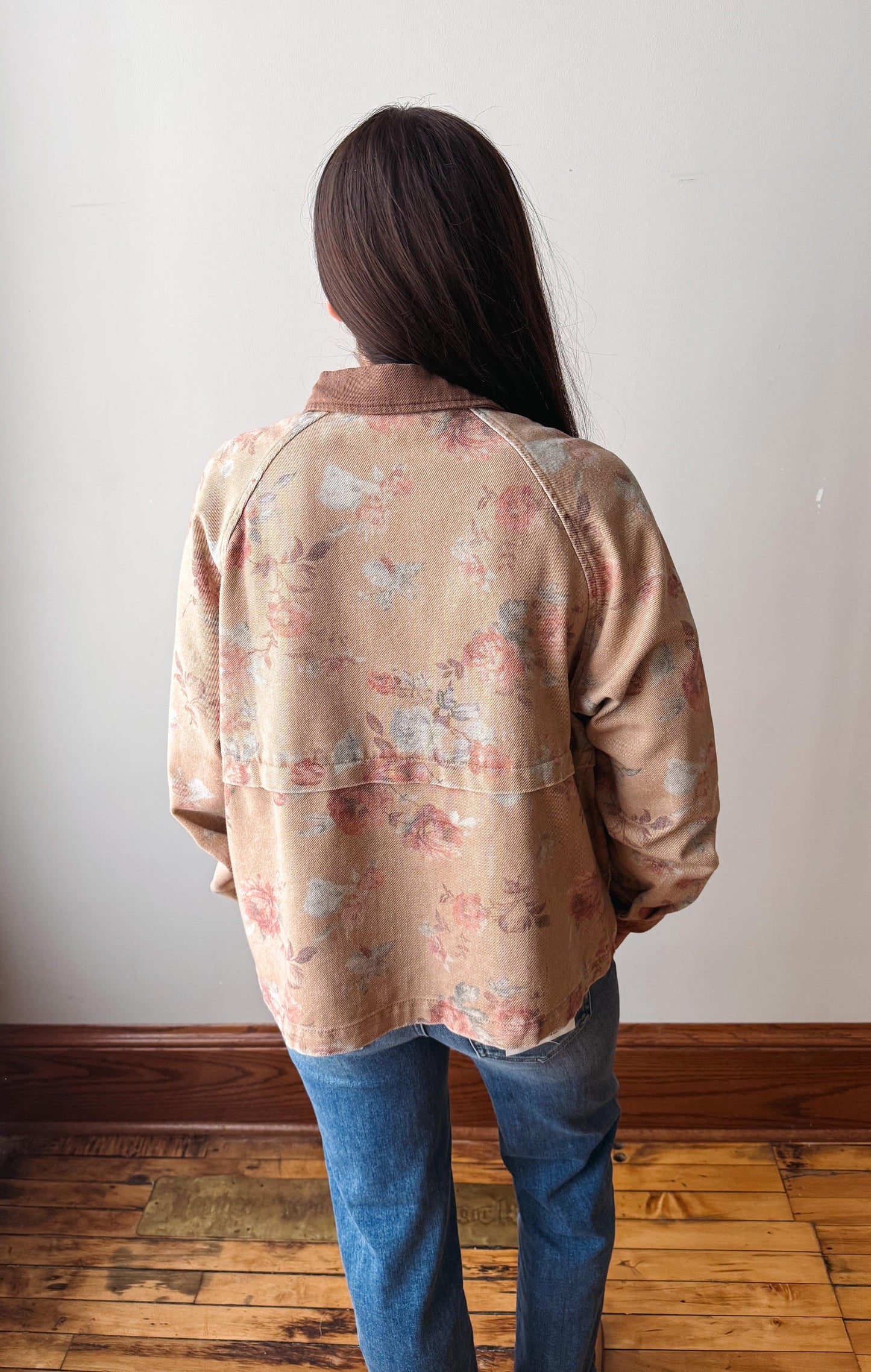 A person viewed from behind wearing a beige floral long-sleeve overshirt with a brown collar and blue jeans, standing on a wooden floor against a plain off-white wall.