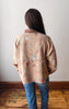 A person viewed from behind wearing a beige floral long-sleeve overshirt with a brown collar and blue jeans, standing on a wooden floor against a plain off-white wall.