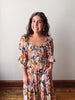 Woman wearing a colorful floral dress standing against a plain wall.