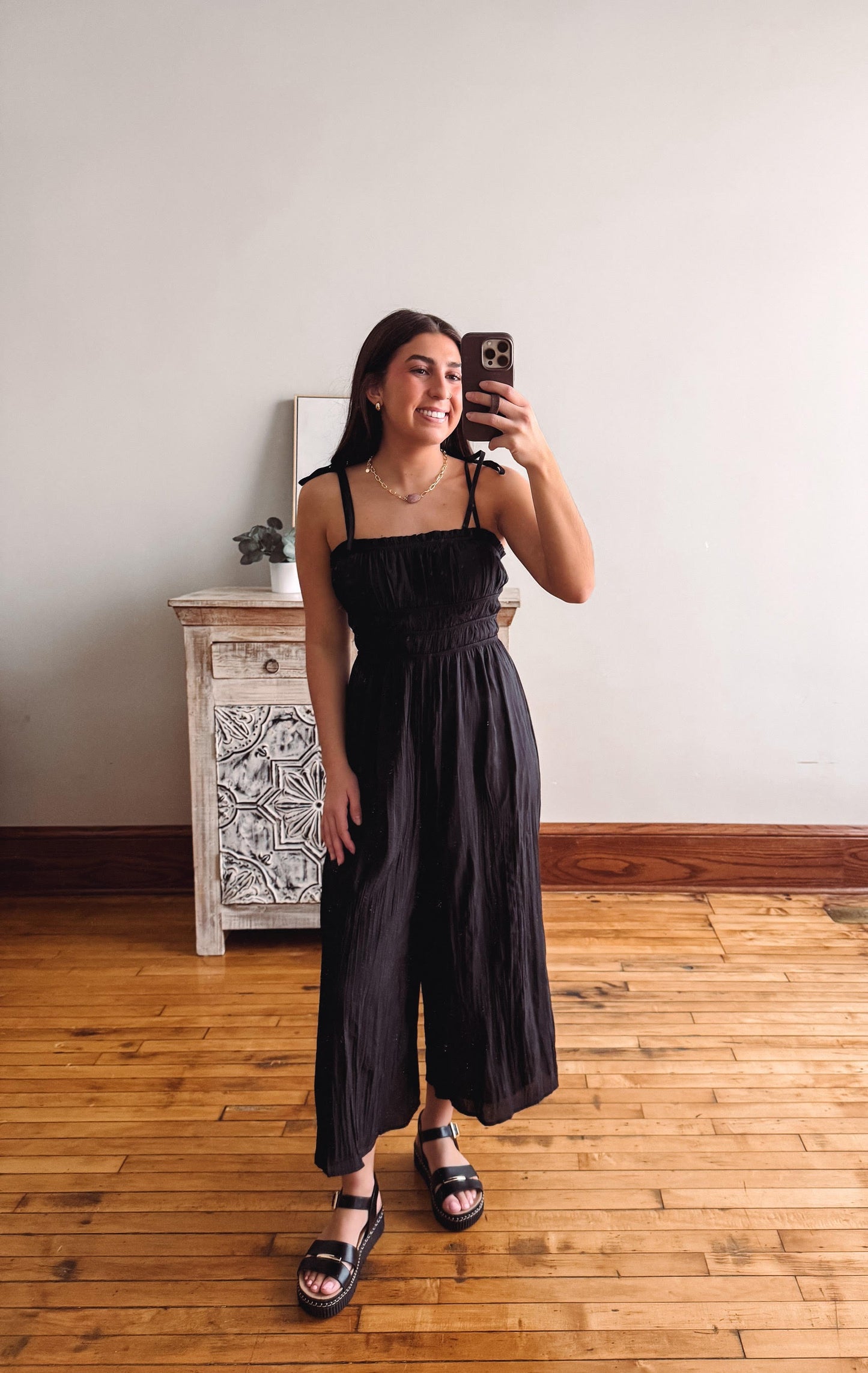 Woman taking a mirror selfie wearing a black dress in a room with wooden flooring and a white wall.