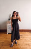 Woman taking a mirror selfie wearing a black dress in a room with wooden flooring and a white wall.