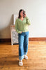 A woman takes a mirror selfie wearing a light sage green knit cardigan, blue cuffed jeans, and white slide sandals in a room with a wooden floor and a white wall.
