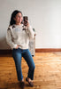 A woman takes a mirror selfie wearing a cream knit sweater with brown leaf patterns and a blue flower motif, paired with blue jeans and brown slip-on shoes, in a room with wooden floors and a white wall.