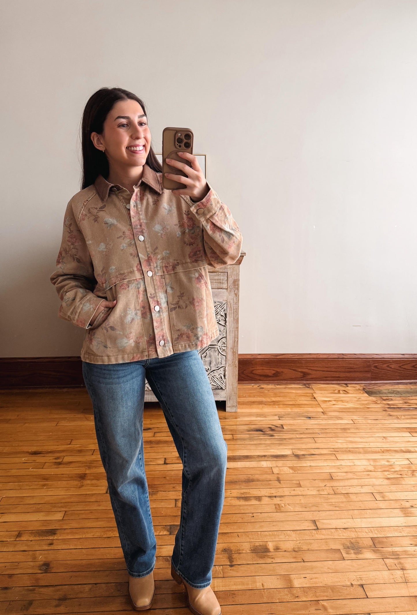A woman takes a mirror selfie wearing a beige button-up shirt with a subtle floral pattern, blue jeans, and tan boots in an indoor room with wooden floors and a plain wall.