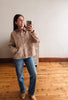A woman takes a mirror selfie wearing a beige button-up shirt with a subtle floral pattern, blue jeans, and tan boots in an indoor room with wooden floors and a plain wall.