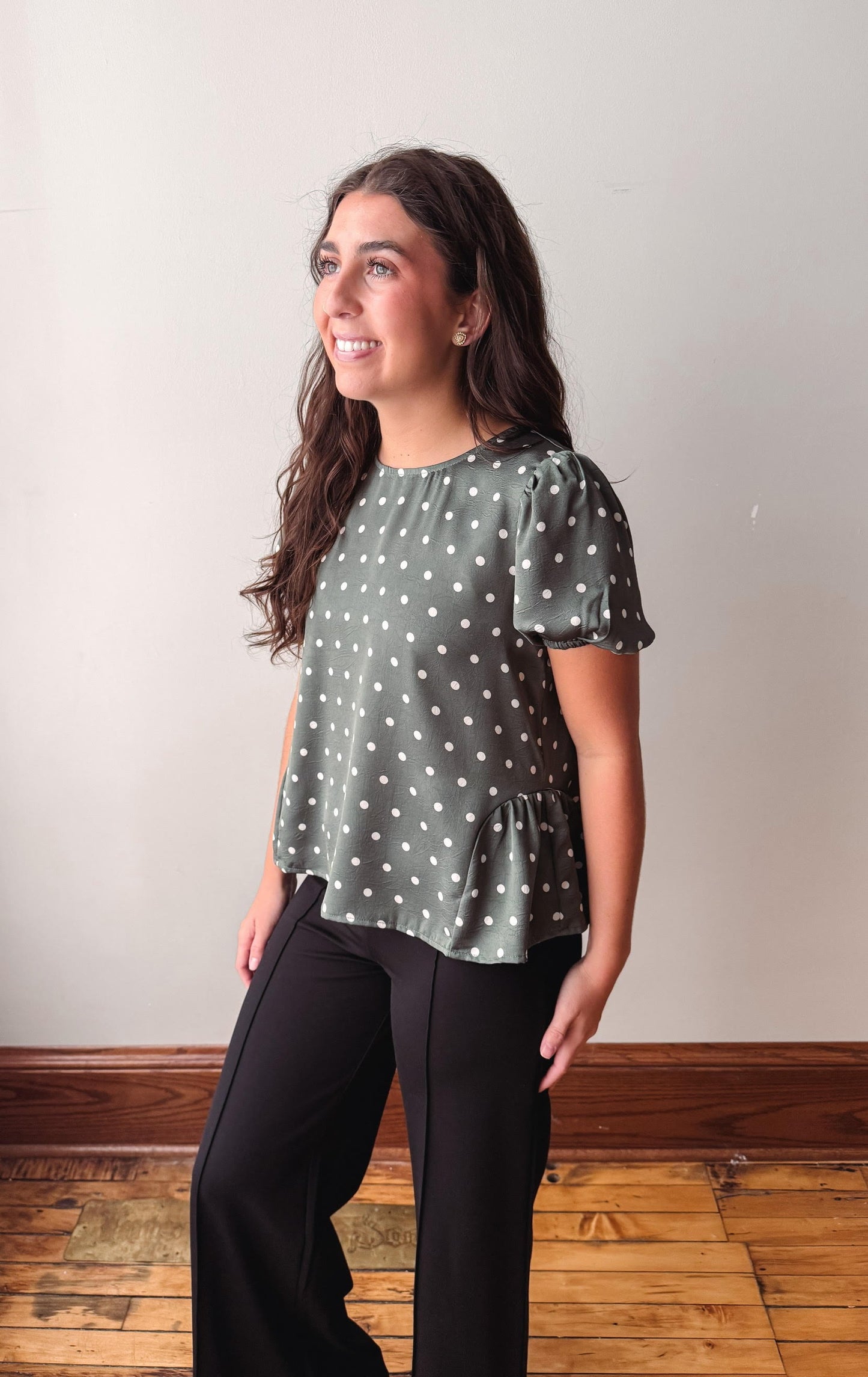 A woman modeling a sage green short-sleeve blouse with white polka dots and black pants, standing in a room with a light gray wall and wooden floor.