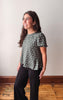 A woman modeling a sage green short-sleeve blouse with white polka dots and black pants, standing in a room with a light gray wall and wooden floor.