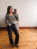 A woman taking a mirror selfie in a room with light-colored walls and a wooden floor, wearing a sage green long-sleeve top, black jeans, and brown boots.