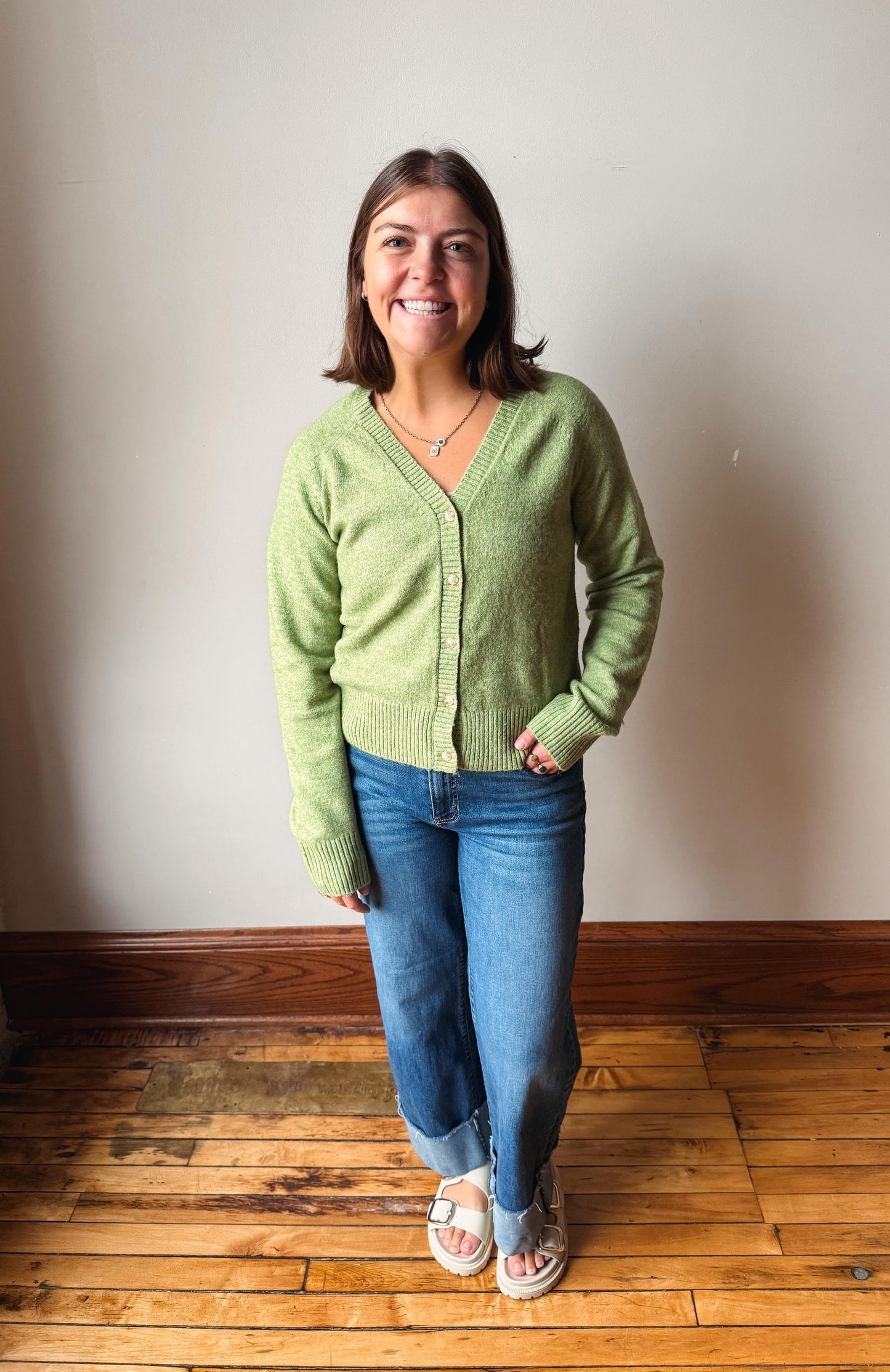 A woman stands in a room with a beige wall and wooden floor, wearing a light green knit cardigan, blue jeans, and white slide sandals.