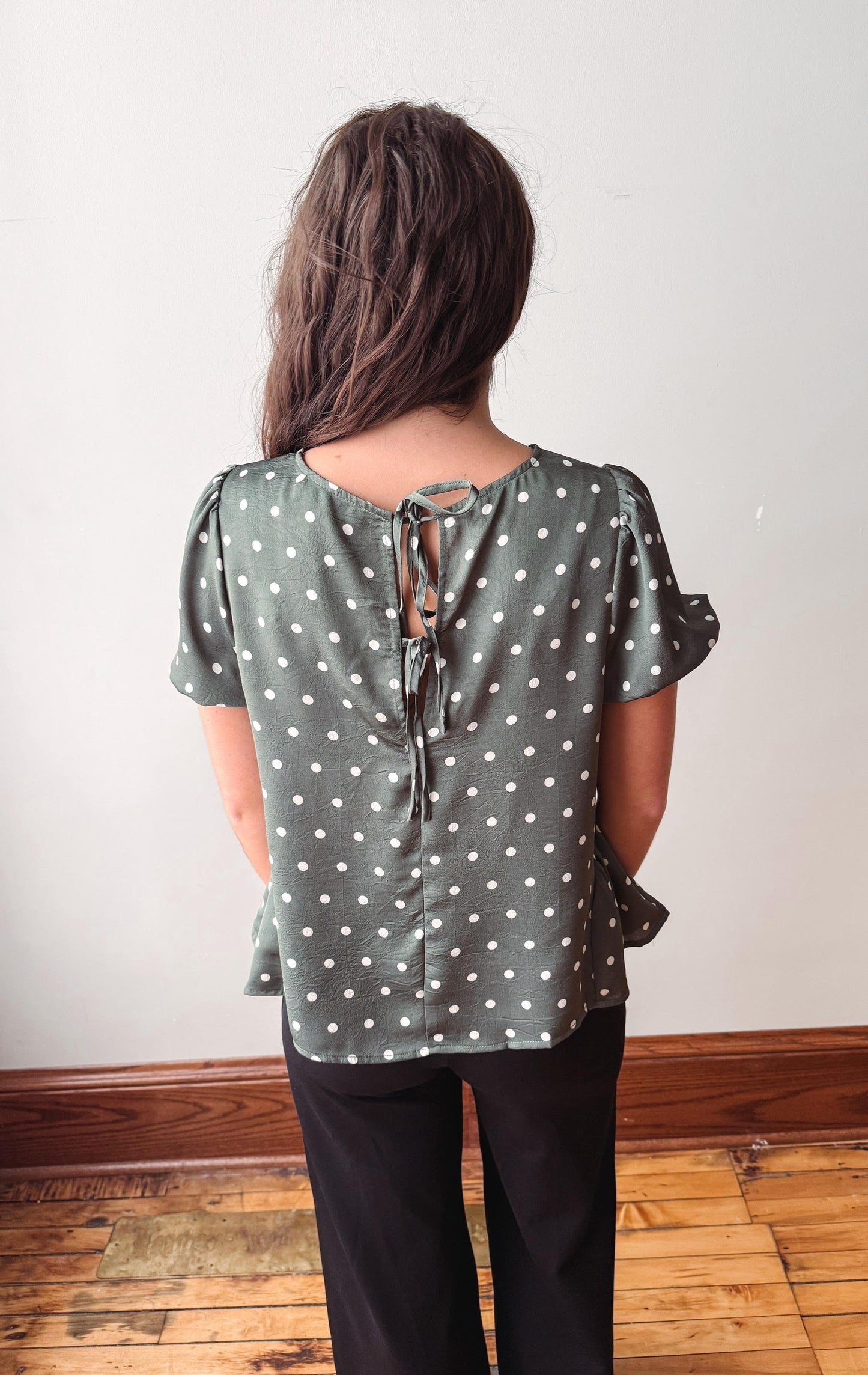A woman viewed from behind wearing a sage green short-sleeve blouse with white polka dots and a tie-back neckline, paired with black pants, standing in an indoor room with a light wall and wooden floor.