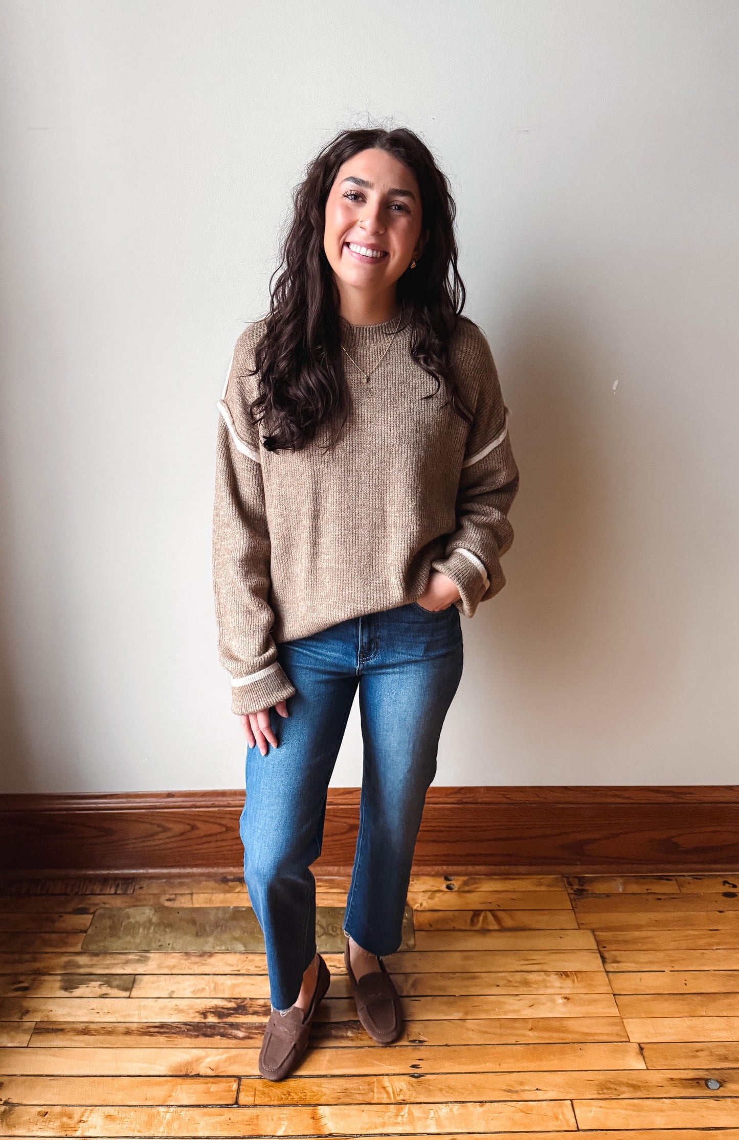 A woman stands in a room with a light-colored wall and wooden floor, wearing a beige knit sweater with white stripes on the sleeves, blue jeans, and brown suede loafers.