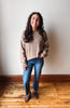 A woman stands in a room with a light-colored wall and wooden floor, wearing a beige knit sweater with white stripes on the sleeves, blue jeans, and brown suede loafers.