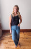 Woman wearing a black sleeveless top and blue jeans standing on a wooden floor.