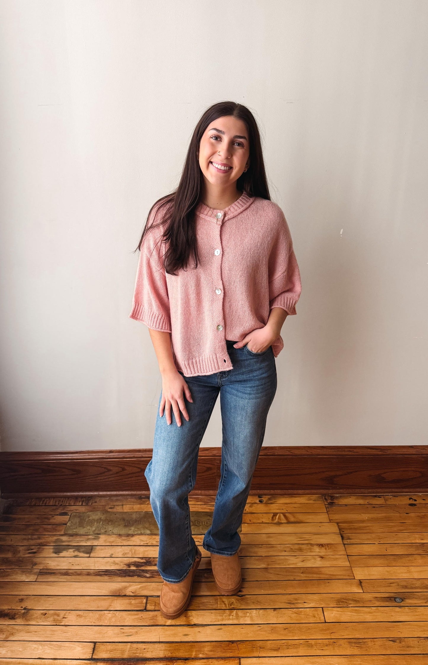 A woman stands in a room with a light beige wall and wooden floor, wearing a pink short-sleeve knit cardigan, blue jeans, and brown boots.