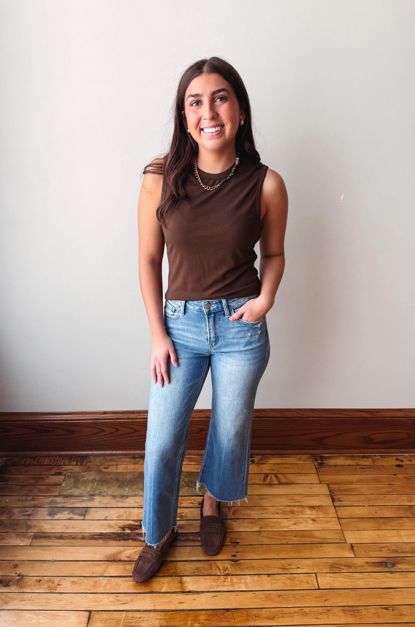 Woman wearing a brown sleeveless top and blue jeans standing on a wooden floor with a white wall background.
