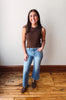 Woman wearing a brown sleeveless top and blue jeans standing on a wooden floor with a white wall background.