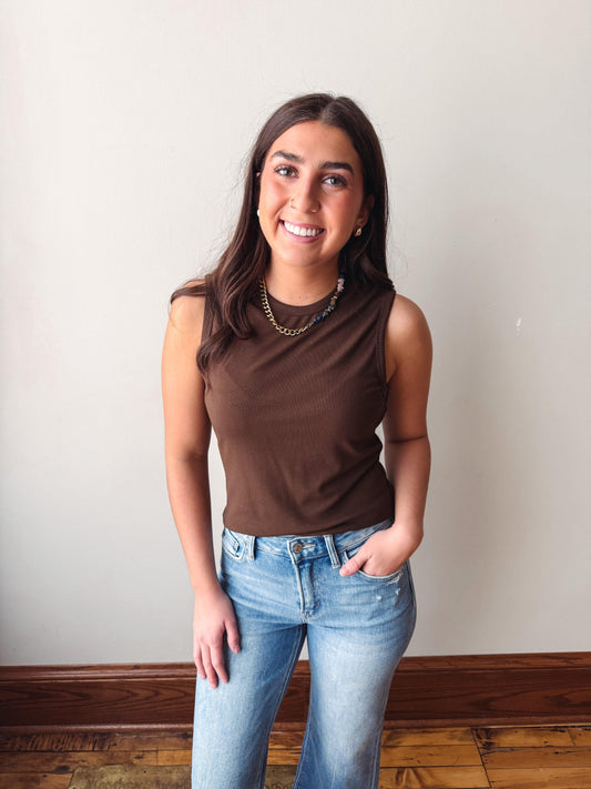 Woman wearing a brown sleeveless top and blue jeans standing against a plain wall.