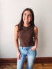 Woman wearing a brown sleeveless top and blue jeans standing against a plain wall.