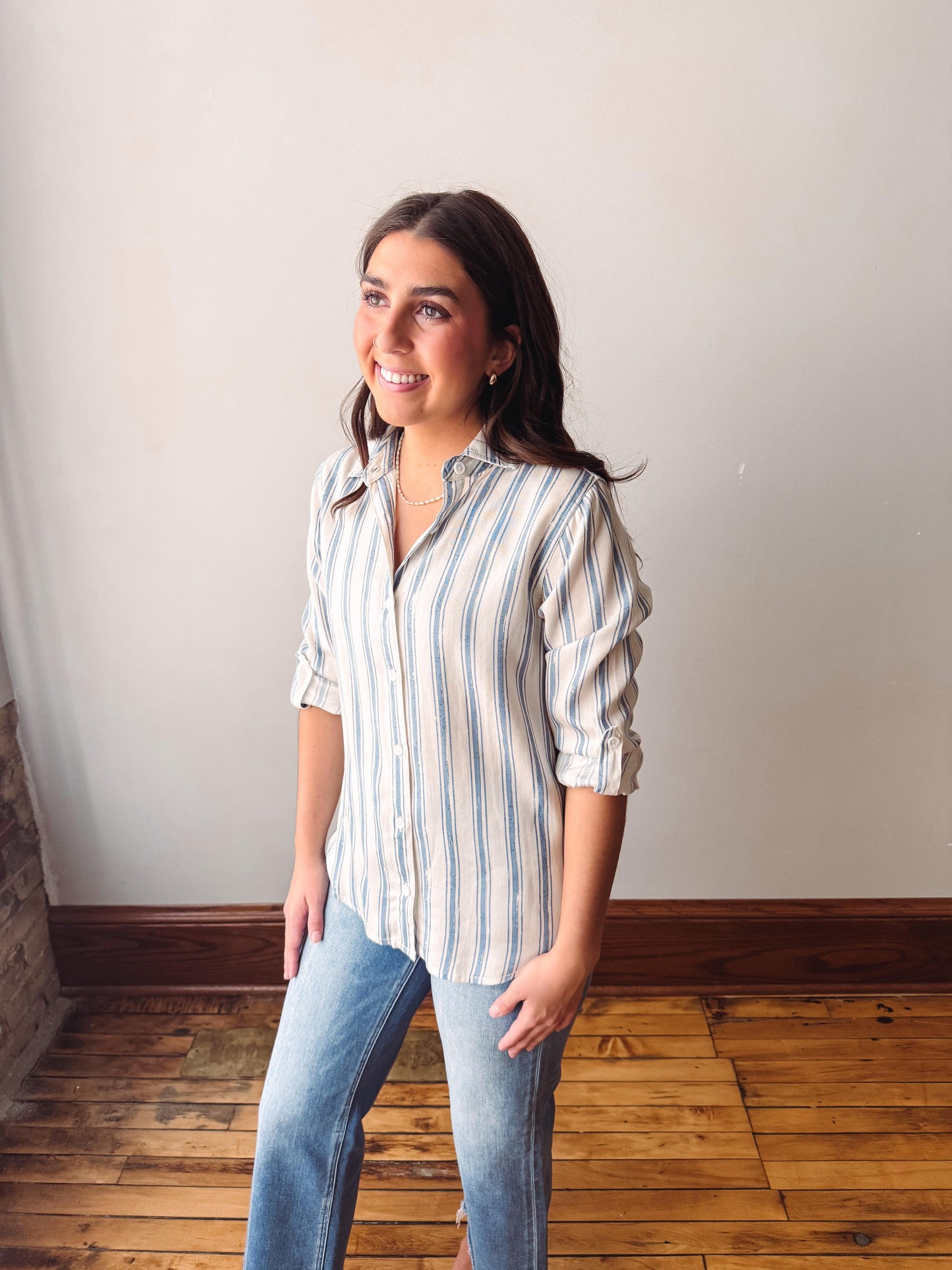 Woman wearing a striped shirt and jeans standing in a room with wooden flooring and white walls.