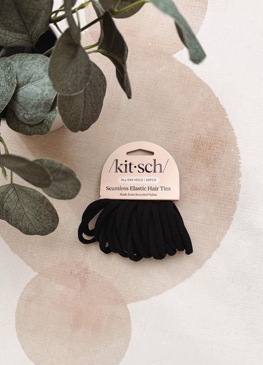Kit-sch seamless elastic hair ties in black, attached to a light pink cardboard hanger, styled on a textured cream background with soft pink circular shapes and green eucalyptus leaves.